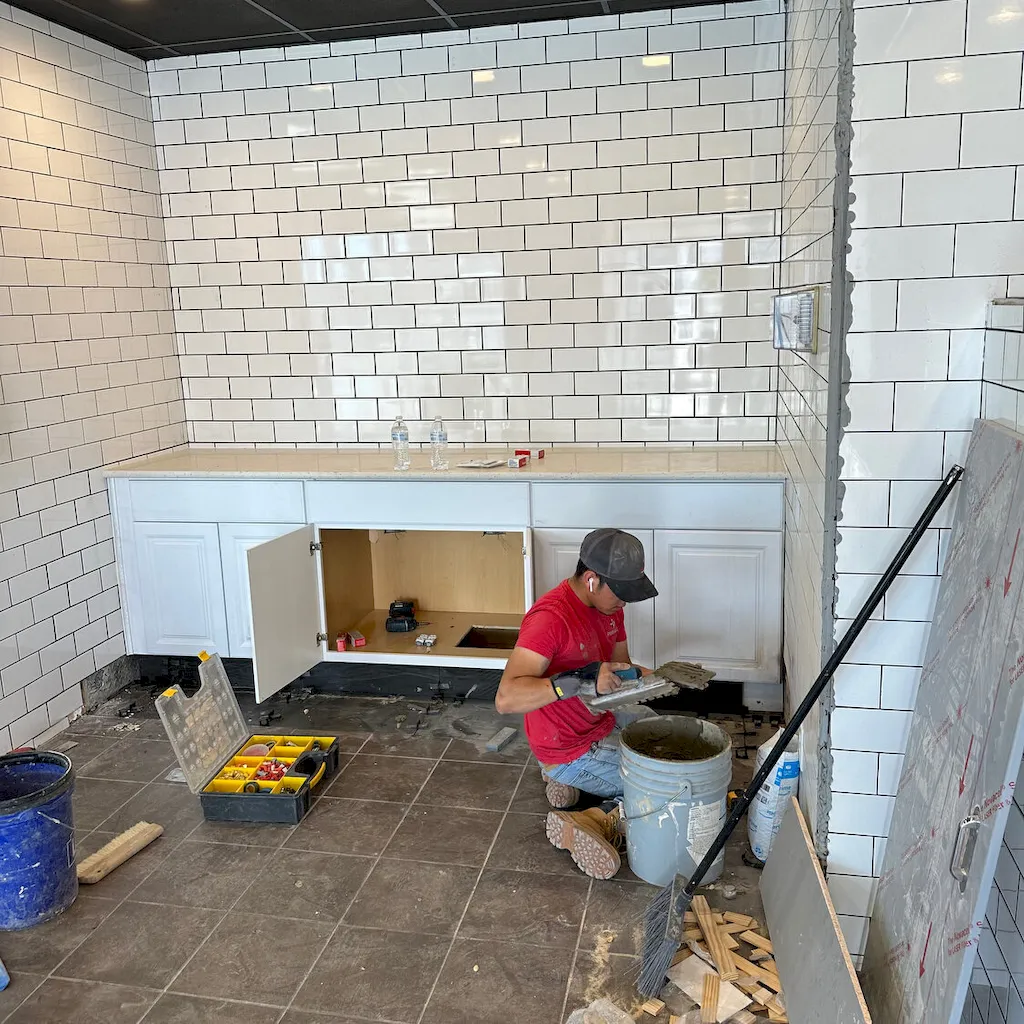 Flooring worker kneeling and mixing tile mortar with a notched trowel in a white subway-tiled room during installation.