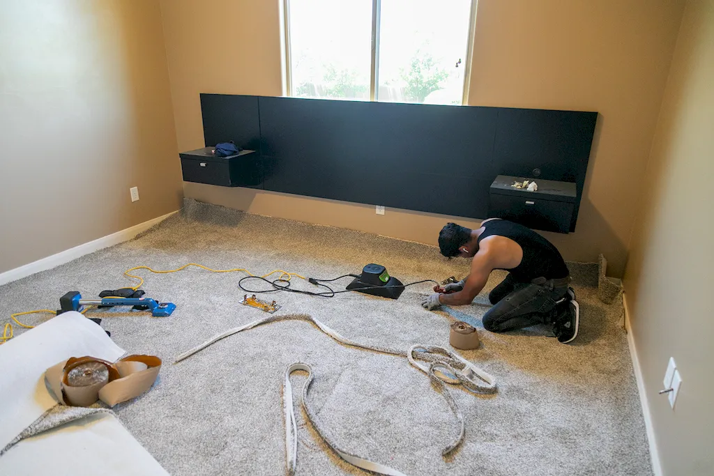 Red Rock Flooring installer kneeling with a seam iron joining bedroom carpet beside a modern black headboard.